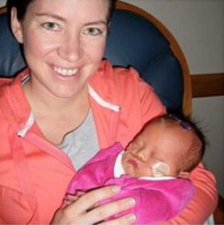 Mother smiles as she sits in chair holding newborn with down syndrome in NICU