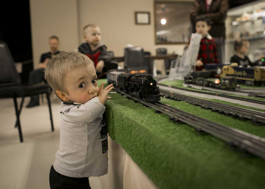 Little boy with dwarfism standing beside table with toy train track on it