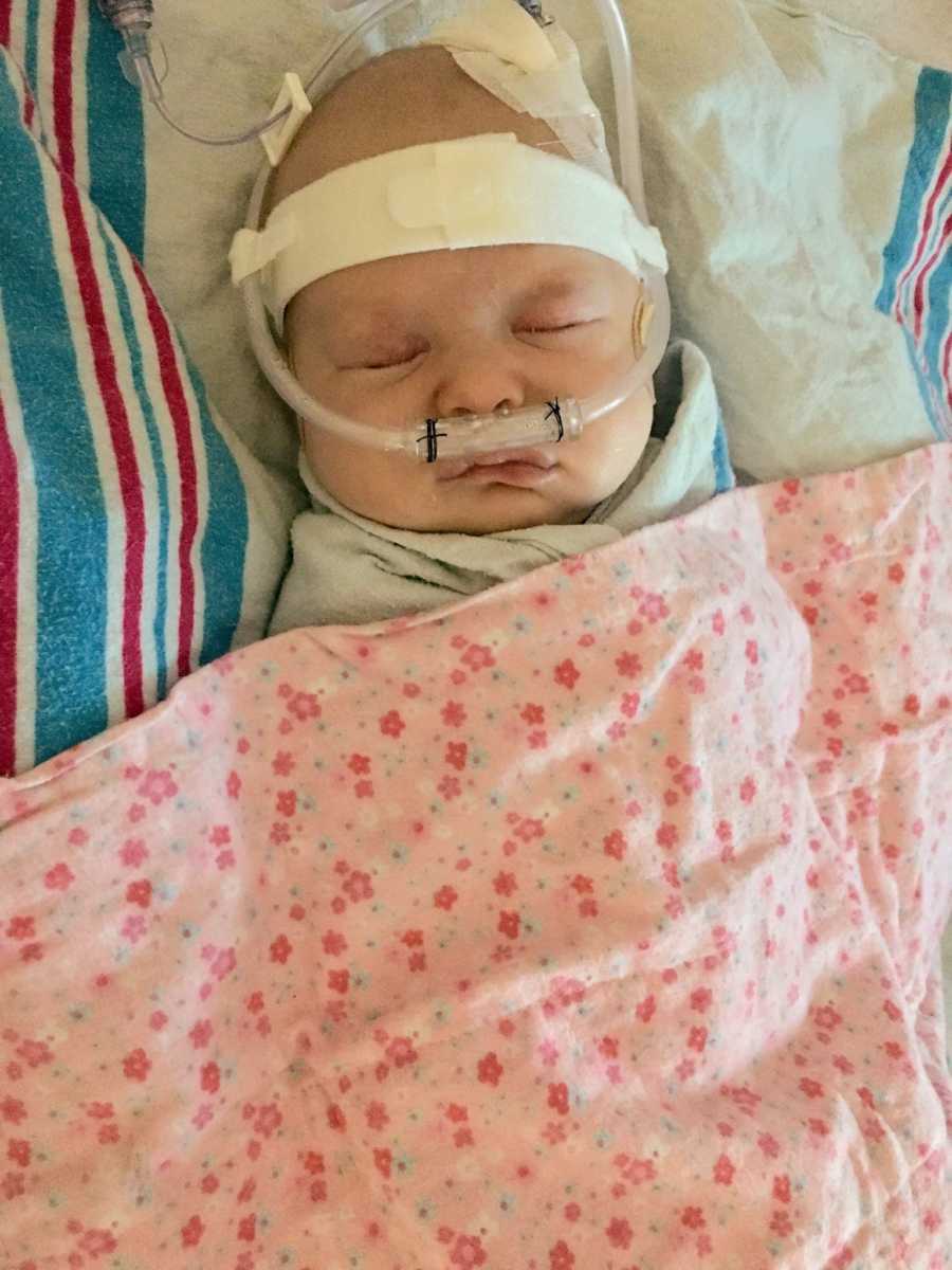 Baby lays in PICU covered in pink floral blanket with oxygen tube up her nose