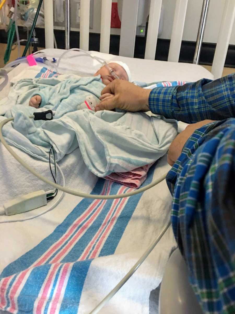Man stands beside baby lying in PICU crib holding her hand