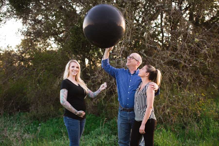 Surrogate stands outside holding stomach while parents of child stands arm in arm looking up at black balloon