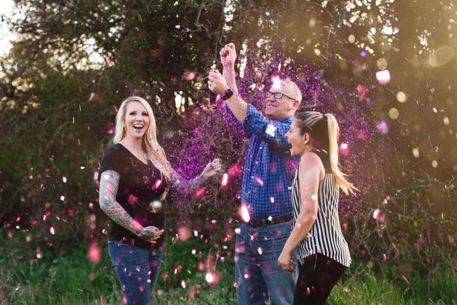 Pregnant surrogate smiling beside parents of child who are covered in pink confetti at gender reveal