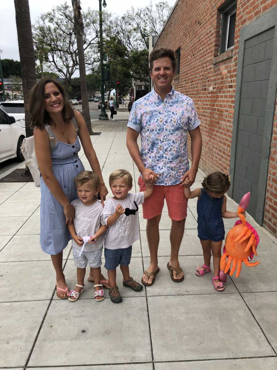 Husband and wife stand on sidewalk holding hands with their toddler triplets