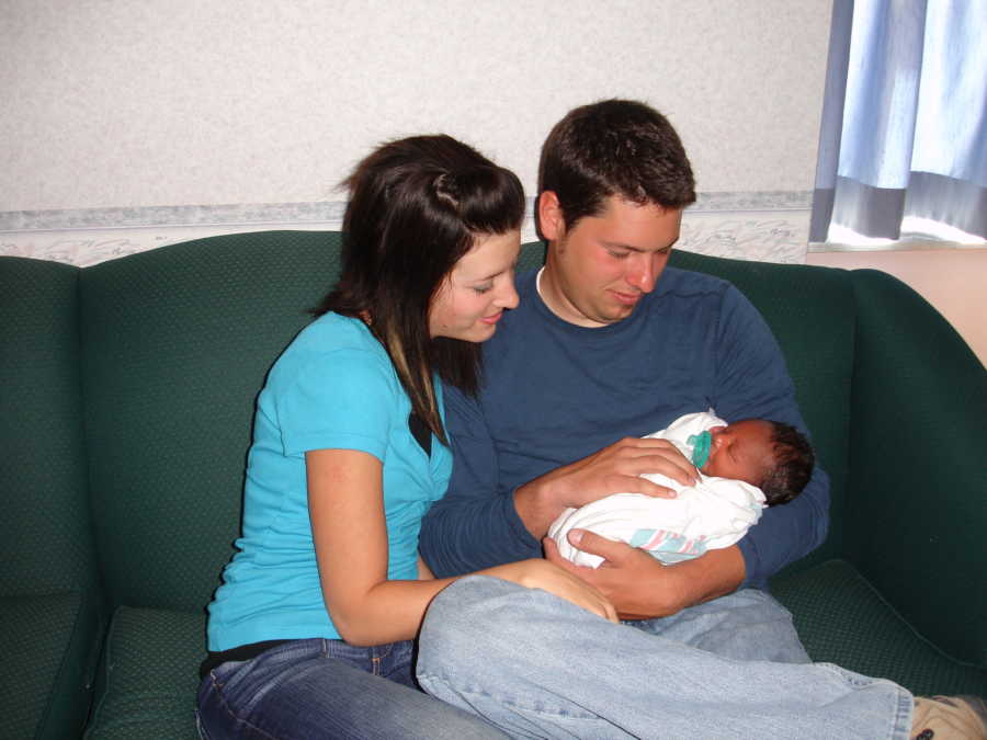 Father sits on couch in hospital holding adopted newborn in his arms with wife sitting beside him