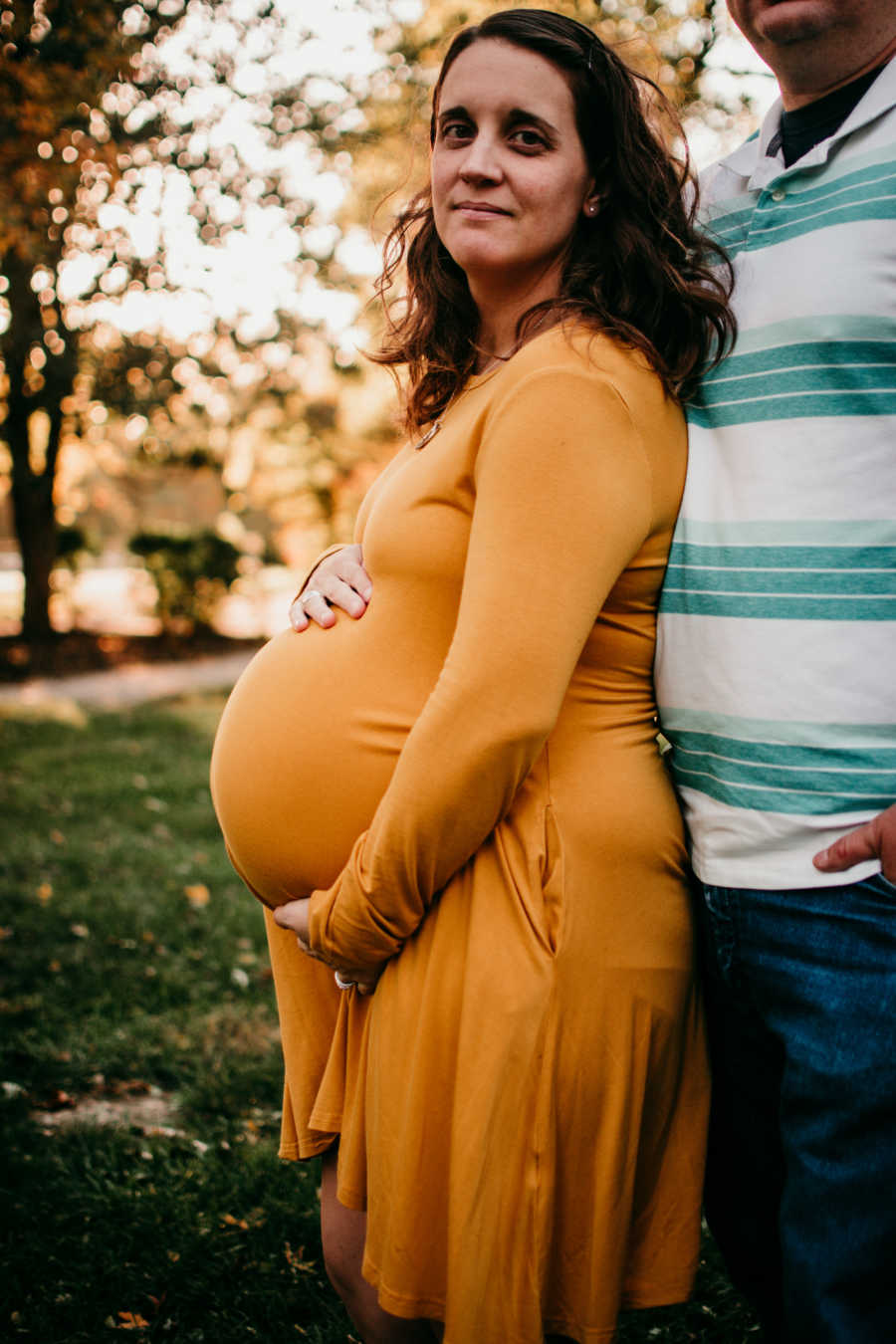 Pregnant birth photographer stands outside holding her stomach beside her husband