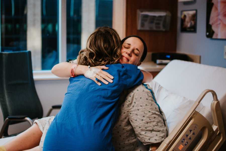 Pregnant birth photographer sits in hospital bed hugging nurse