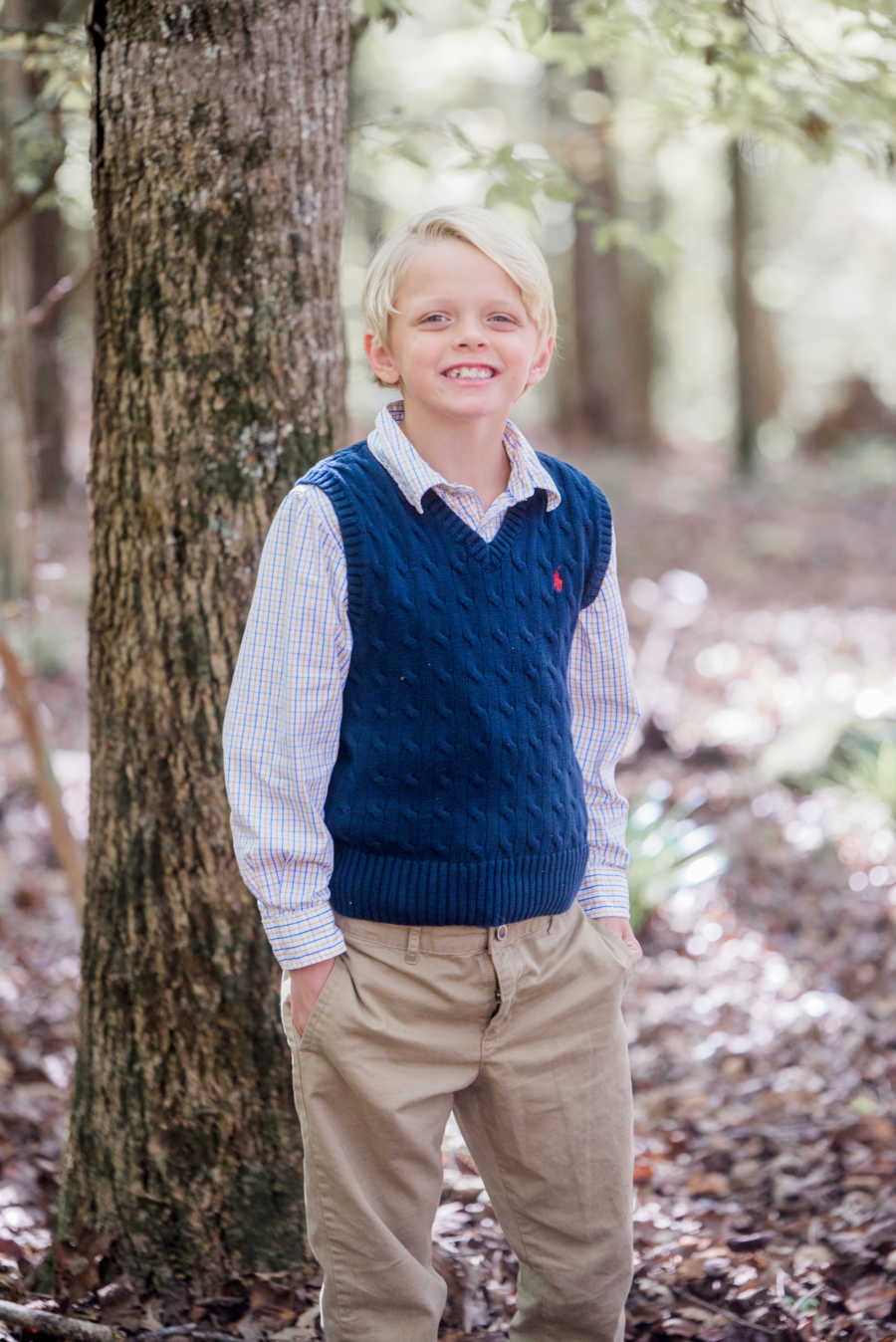 Young boy who told his mom he's "bad" stands in khakis, button down, and sweater vest beside tree