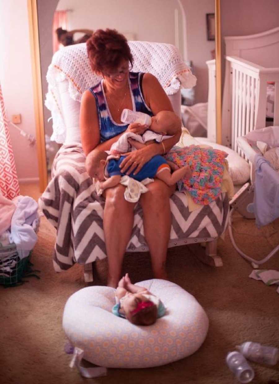 Grandmother sitting in rocking chair feeding one baby and burping another while third baby sleeps on pillow