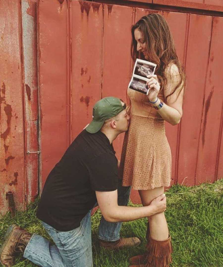 Pregnant woman stands outside holding ultrasound pictures while her husband kisses her stomach