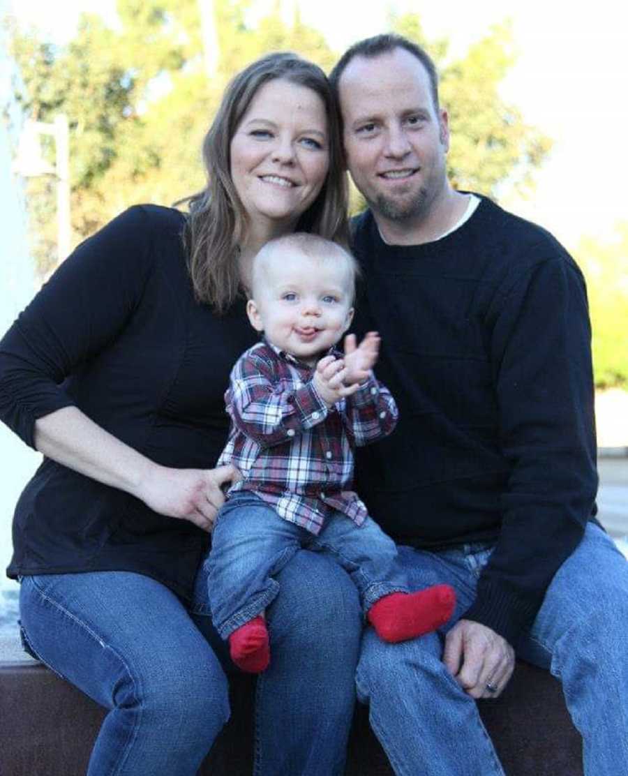 Husband and wife smile outside as their baby son sits on their lap