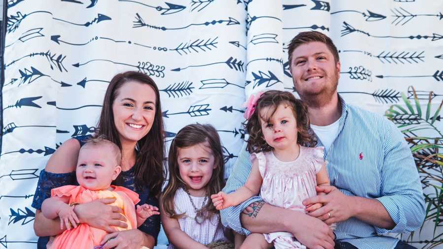 Husband and wife sit in front of backdrop with their adopted daughter and two biological daughters