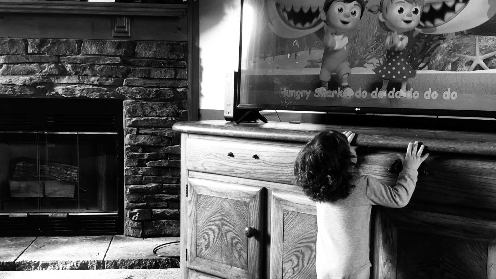 Foster baby stands beside tv stand looking up at tv