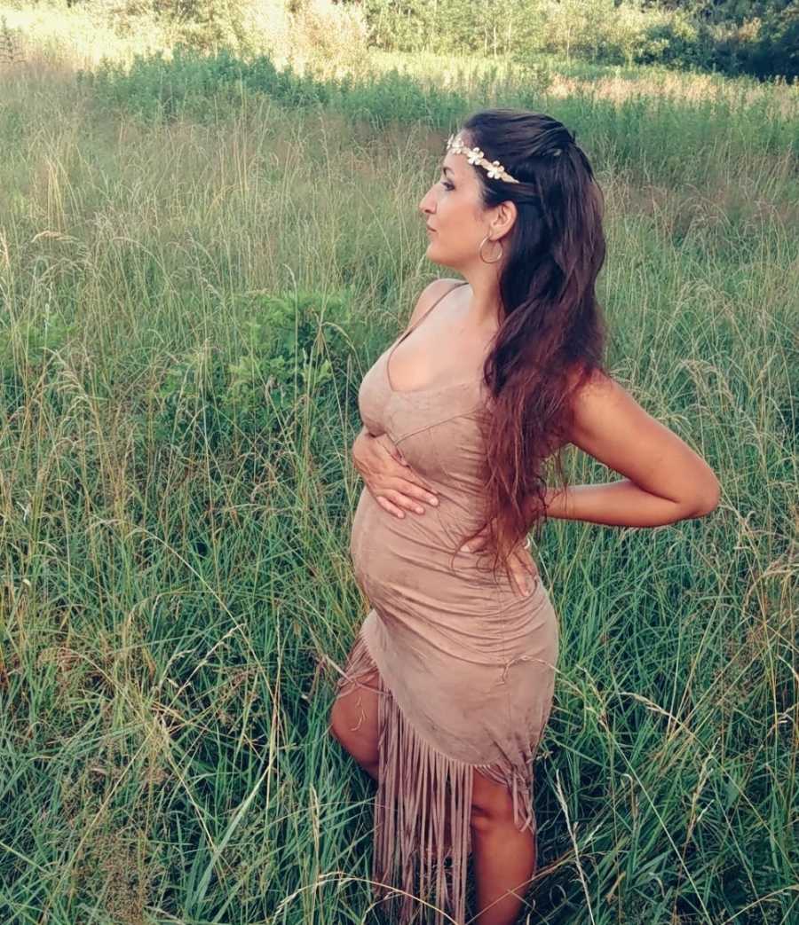 Pregnant woman stands in field in suede fringe dress holding her stomach