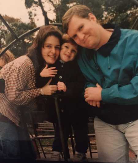 Husband and wife smile beside wife's child on play structure