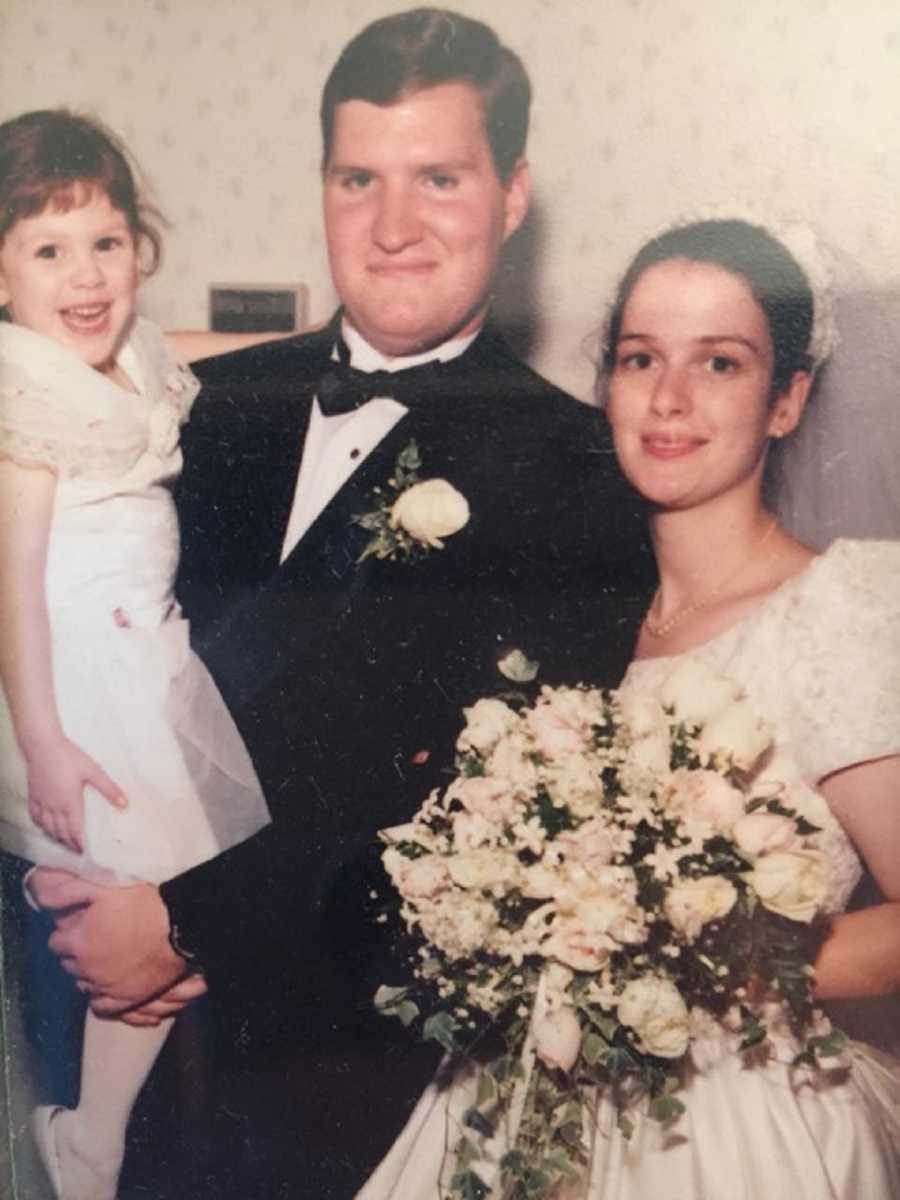 Bride and groom stand smiling while groom holds brides daughter