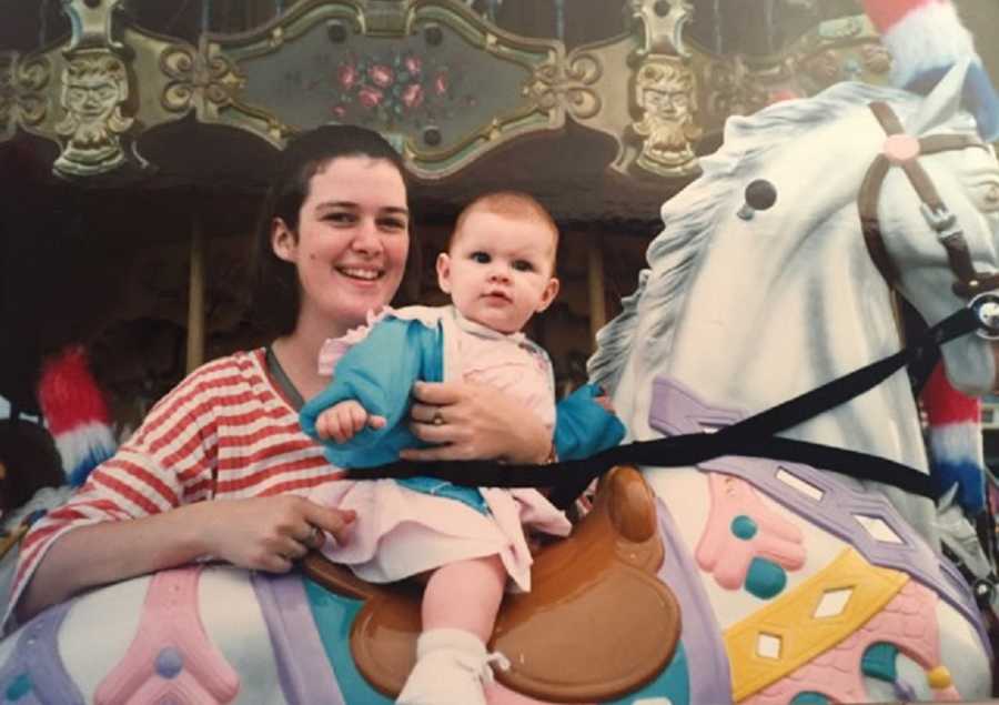 Mother smiles as she holds daughter that is sitting on horse on carousel