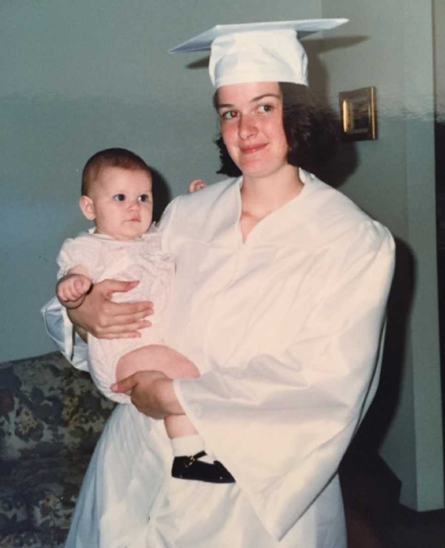Woman standing in white cap in gown holds baby daughter she had in high school