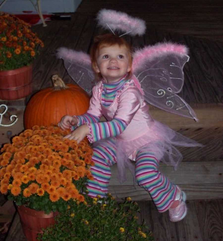 Little girl in pink angel Halloween costume sits on steps outside of home beside pumpkin and orange flowers