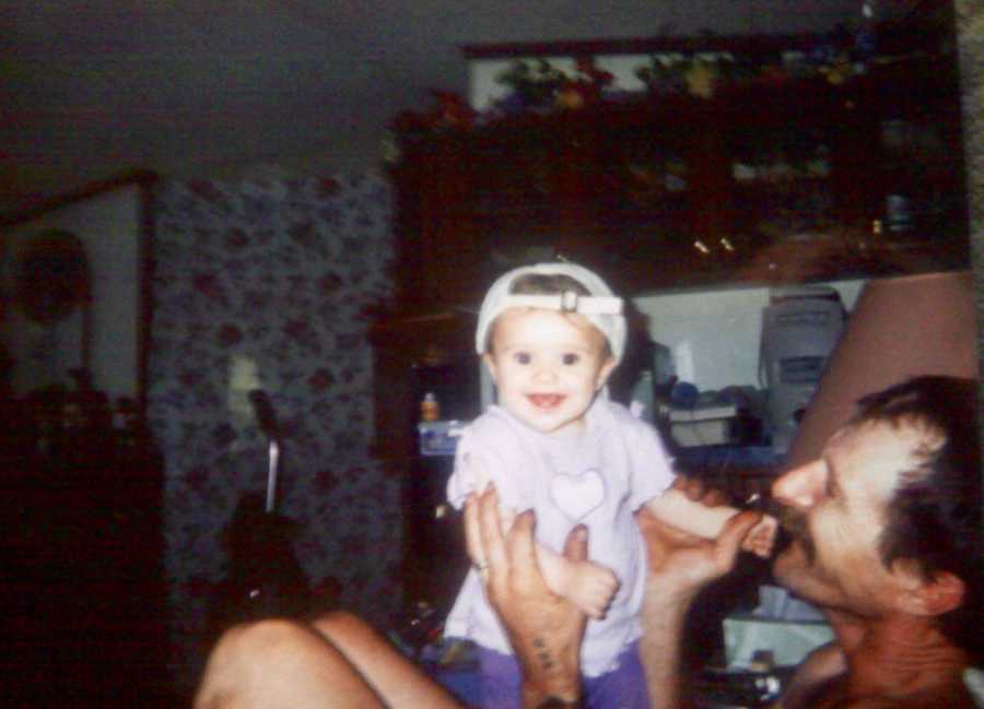 Baby girl with backwards hat on smiles as she stands on father's lap as he smiles at her