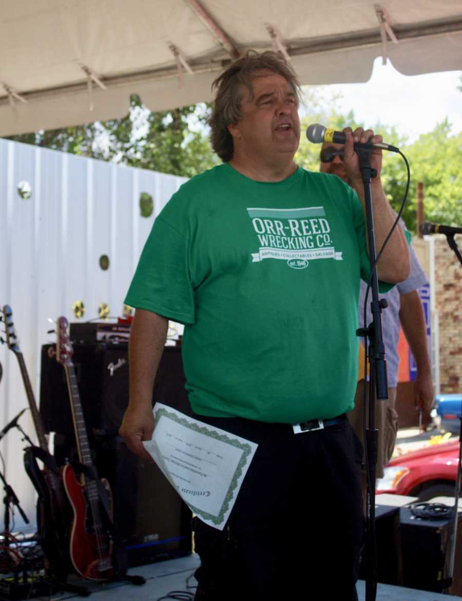 Man who has since passed stands on outdoor stage speaking into microphone