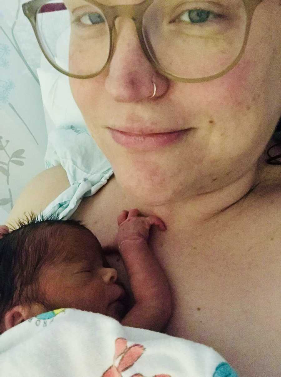 Mother smiles in selfie while newborn baby lays asleep on her chest