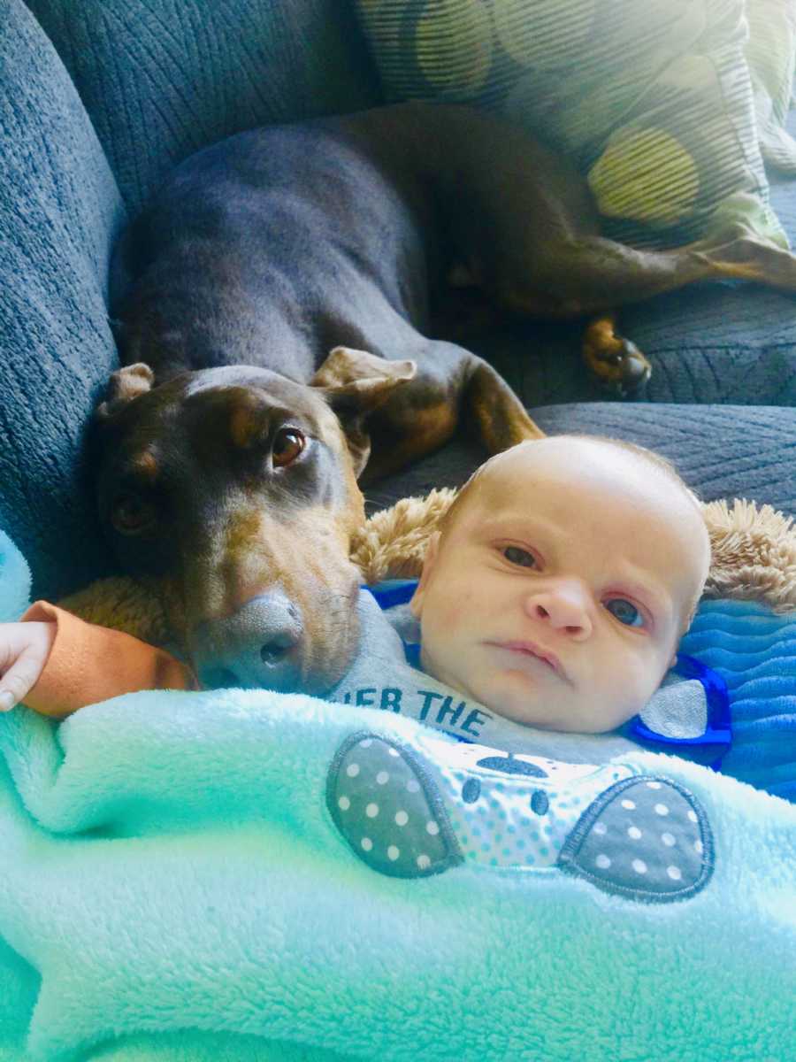 Baby boy lays on his back on couch while dog rests his head on the baby's shoulder