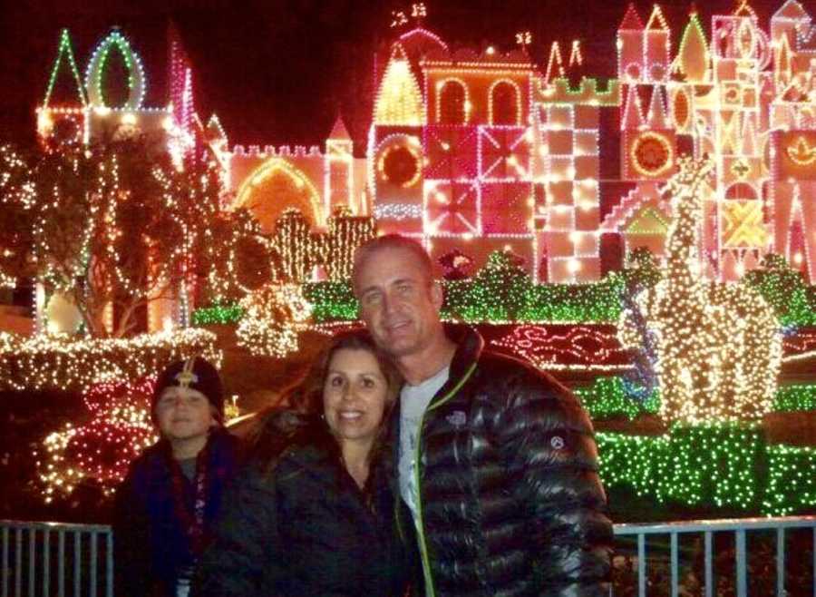 Husband and wife stand smiling arm in arm with their son and Christmas light show behind them