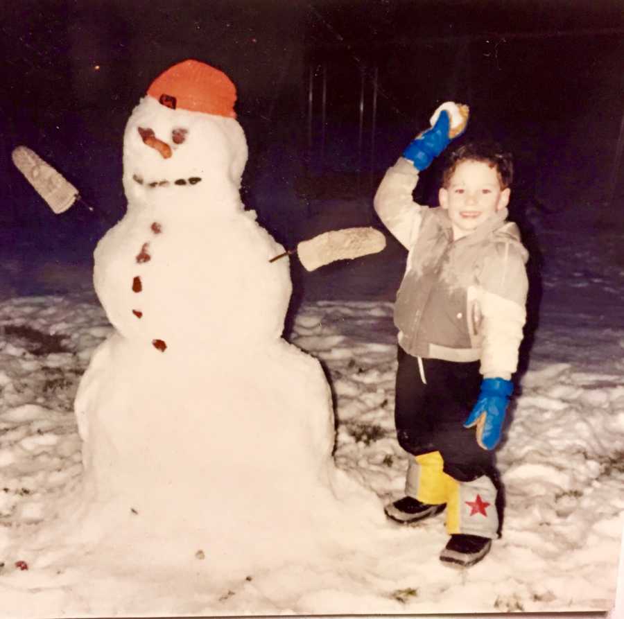 Little boy standing outside beside snowman holding snowball