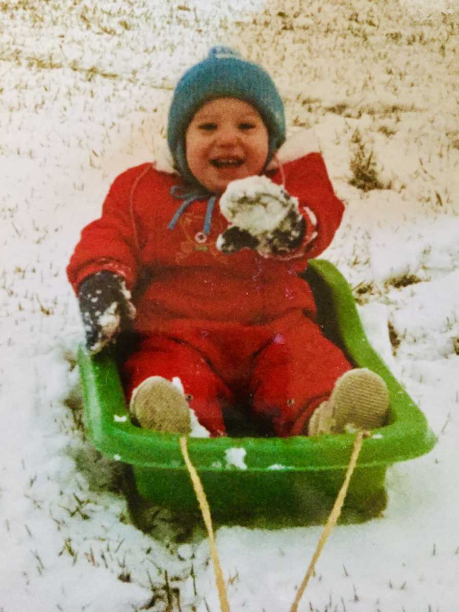 Little kid sitting in green sled smiling with snow suit on