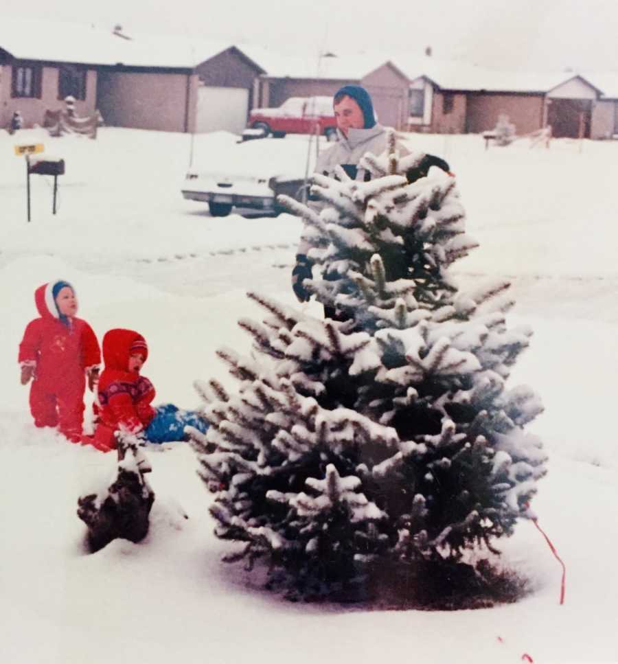 Little kids sitting in yard with dad beside snow covered balsam tree