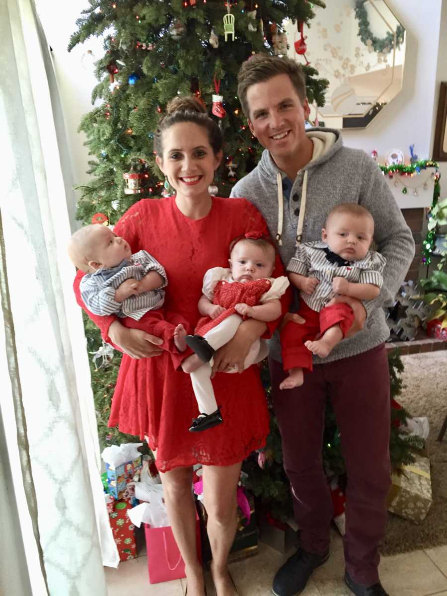 Husband and wife stand in front of Christmas tree in home holding baby triplets