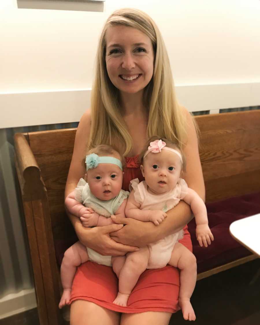 Mother smiles as she sits with twin girls with down syndrome on her lap