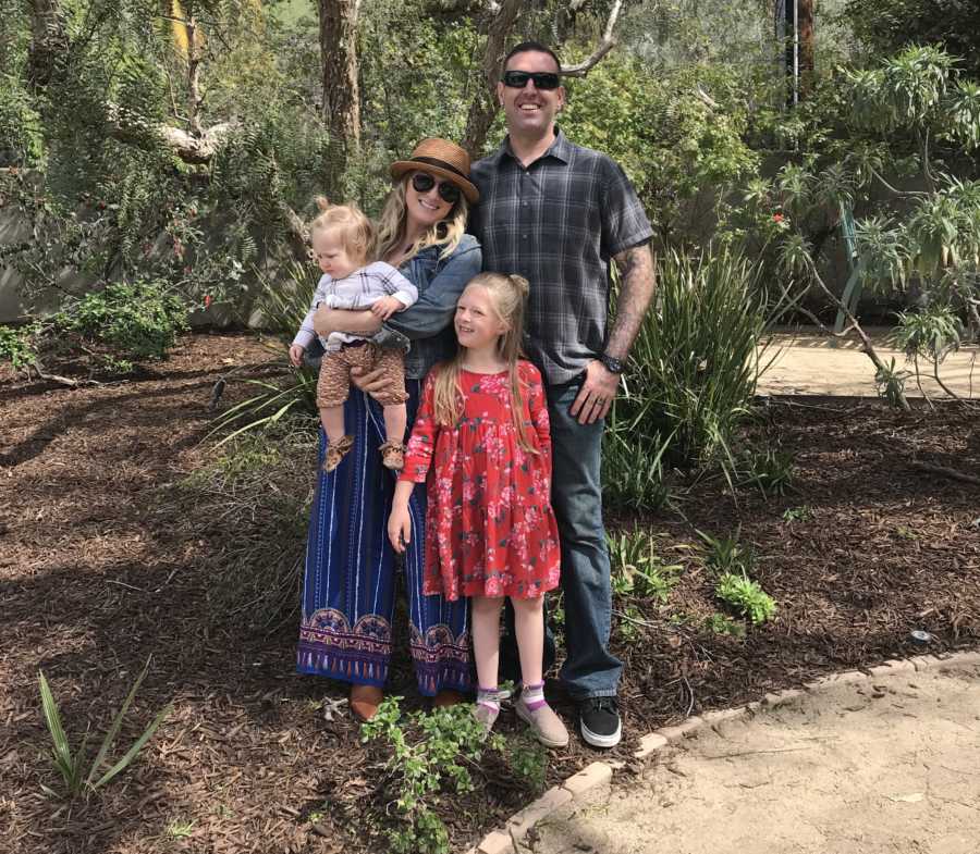 Sober mother stands holding baby beside husband and daughter besides plants