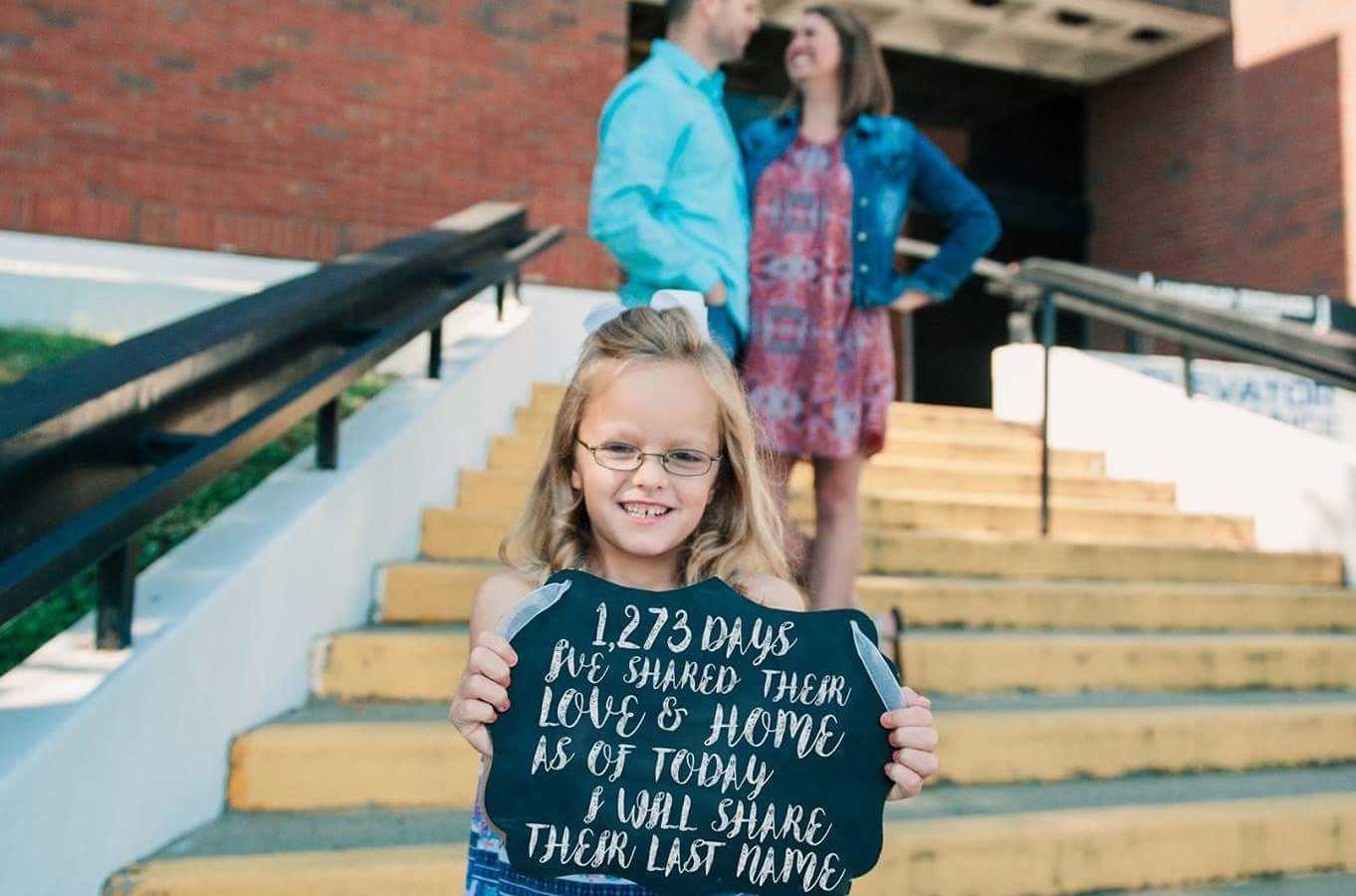 Little girl stands on steps holding sign saying "...as of today I will share their last name" with adopted parents standing behind her