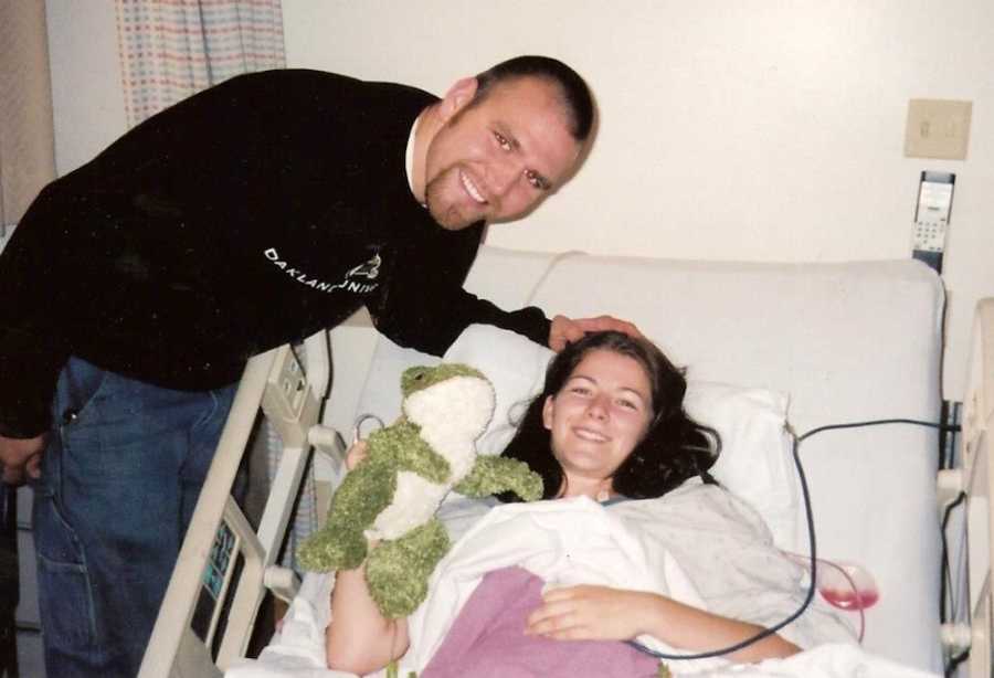 Woman with breast cancer lays in hospital bed holding up frog stuffed animal as fiancee leans over her