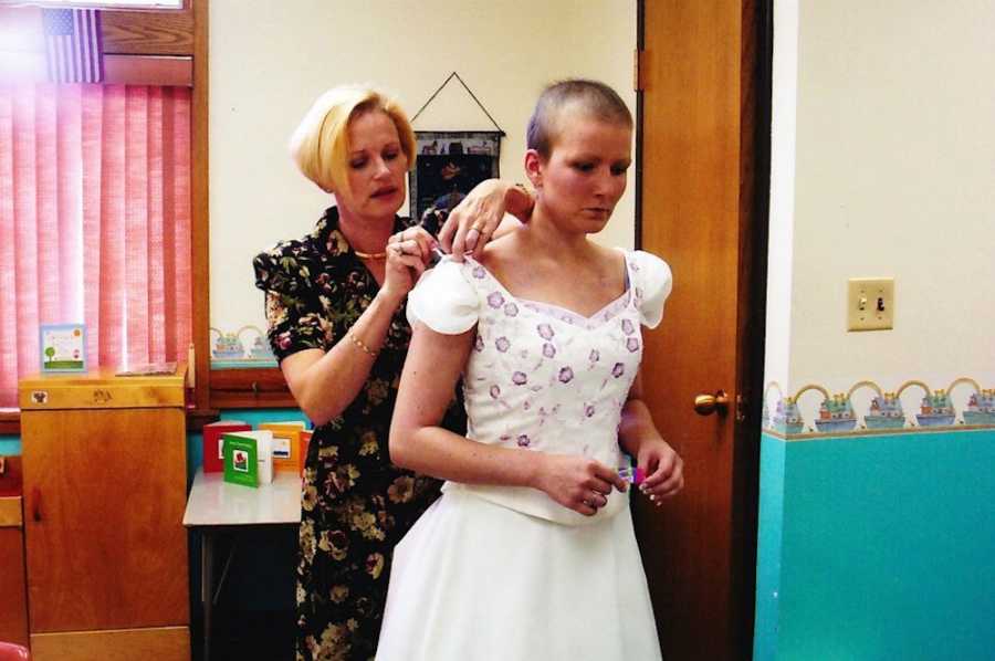 Woman stands behind bride with breast cancer buttoning her dress in hospital room