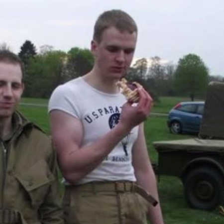 Man wearing tight shirt and cargo pants stands outside holding food to his face