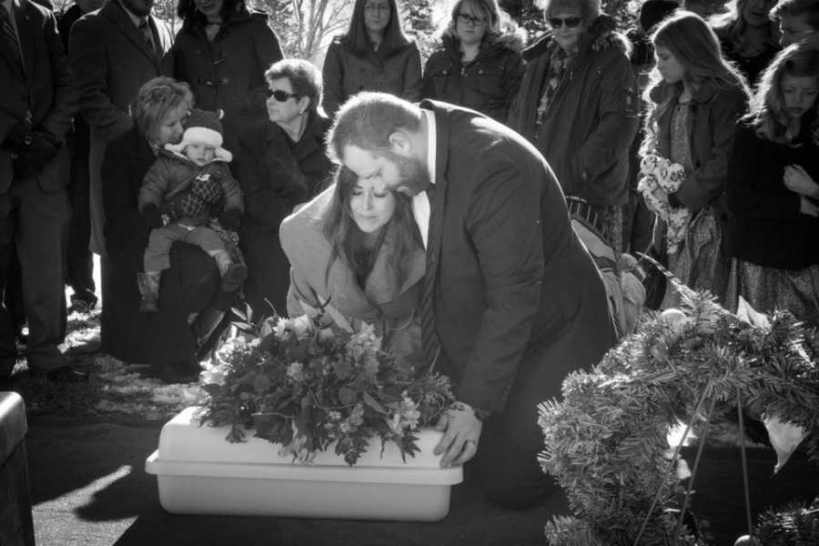 Husband and wife kneel of newborn's grave at funeral with crowd behind them