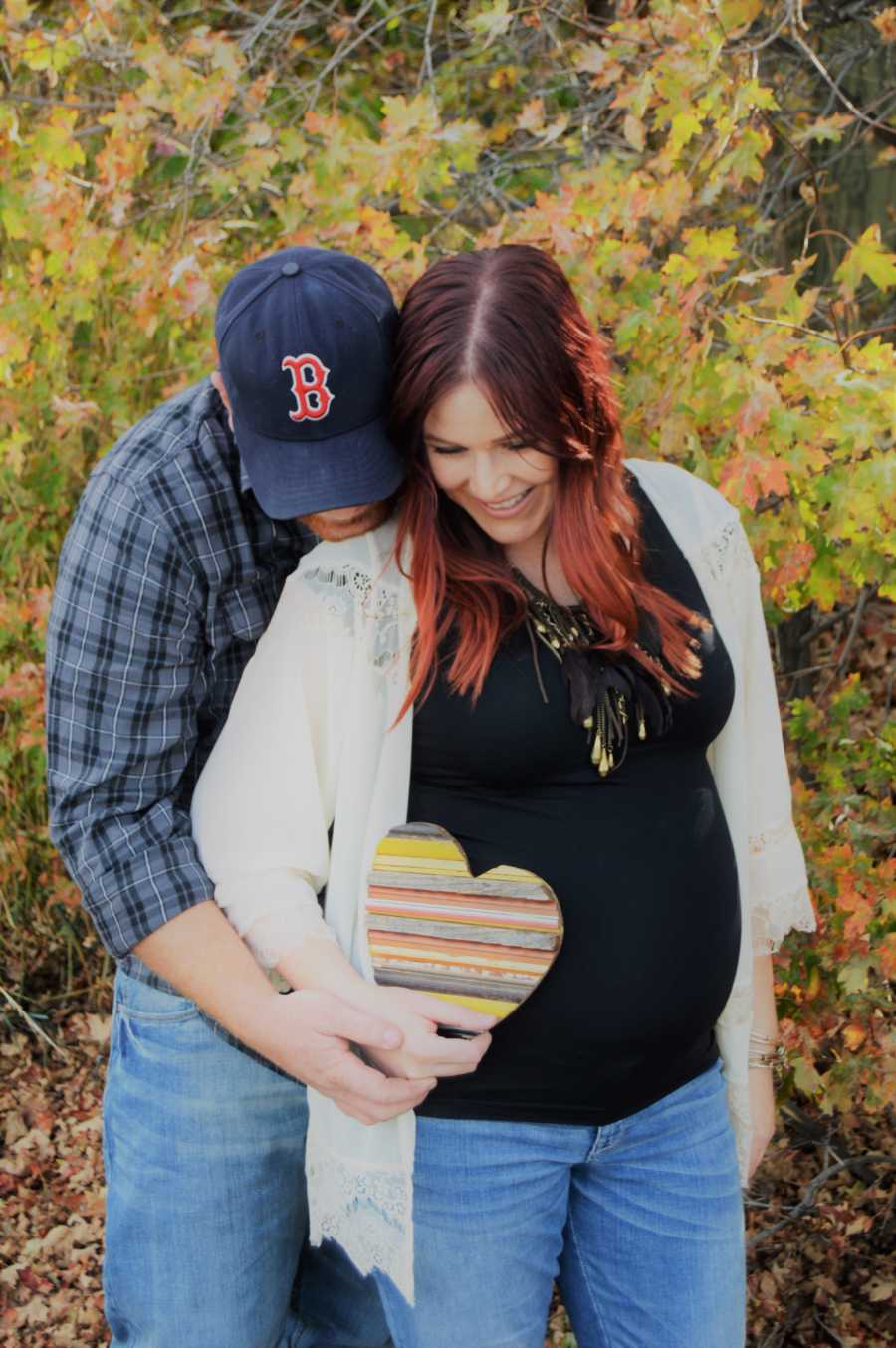 Pregnant wife smiles as she holds wooden heart and husband stands behind her with chin on her shoulder