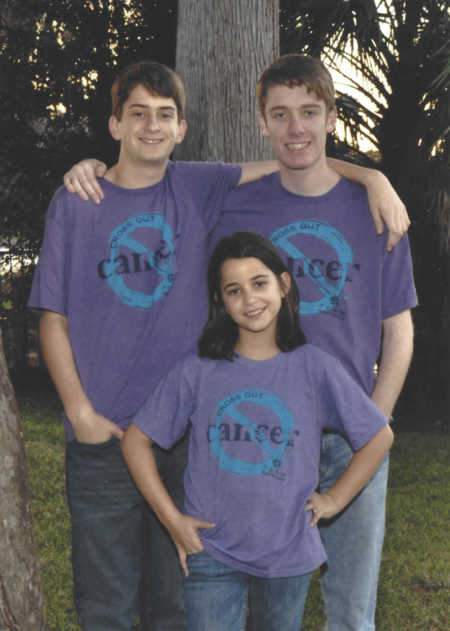 Teen boys stand outside with arms around each other while their sister stands in front of them in matching t-shirts