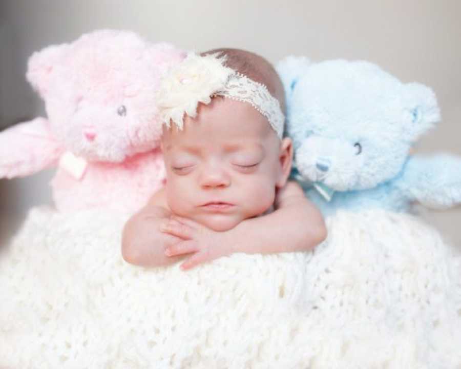 Baby girl who passed away rests her head on her arms as she sits beside pink and blue stuffed bear