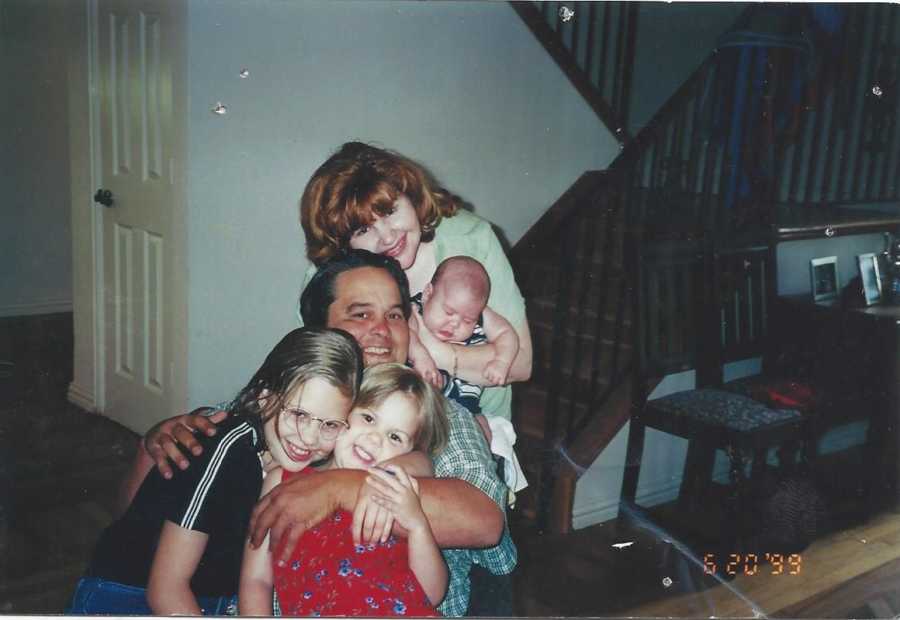 Father who is now deceased smiles in home holding arm around two daughter with wife holding baby behind him