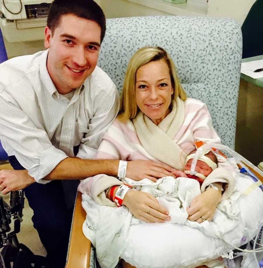 Mother sits in hospital chair with newborn who needs open heart surgery in her lap with husband sitting beside her