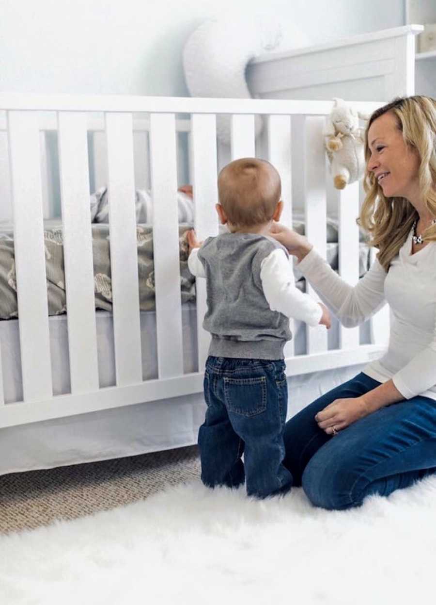 Mother kneels on floor beside young son who had open heart surgery who stands beside his crib