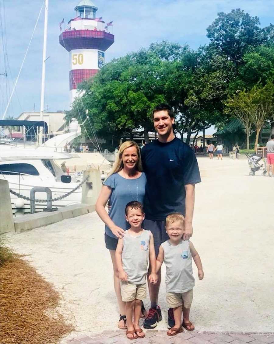 Husband and wife stand with their two sons near marina with lighthouse in background