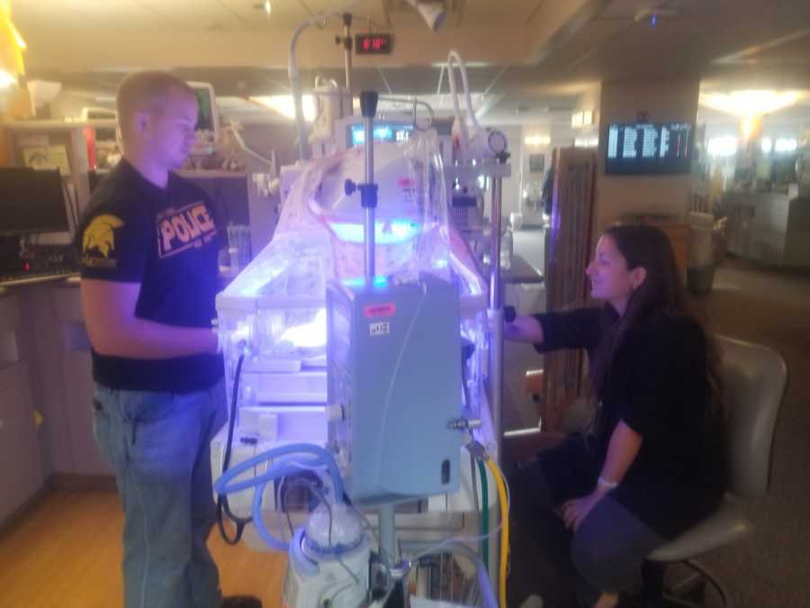 Mother sits while father stands looking at baby laying under blue lights in NICU