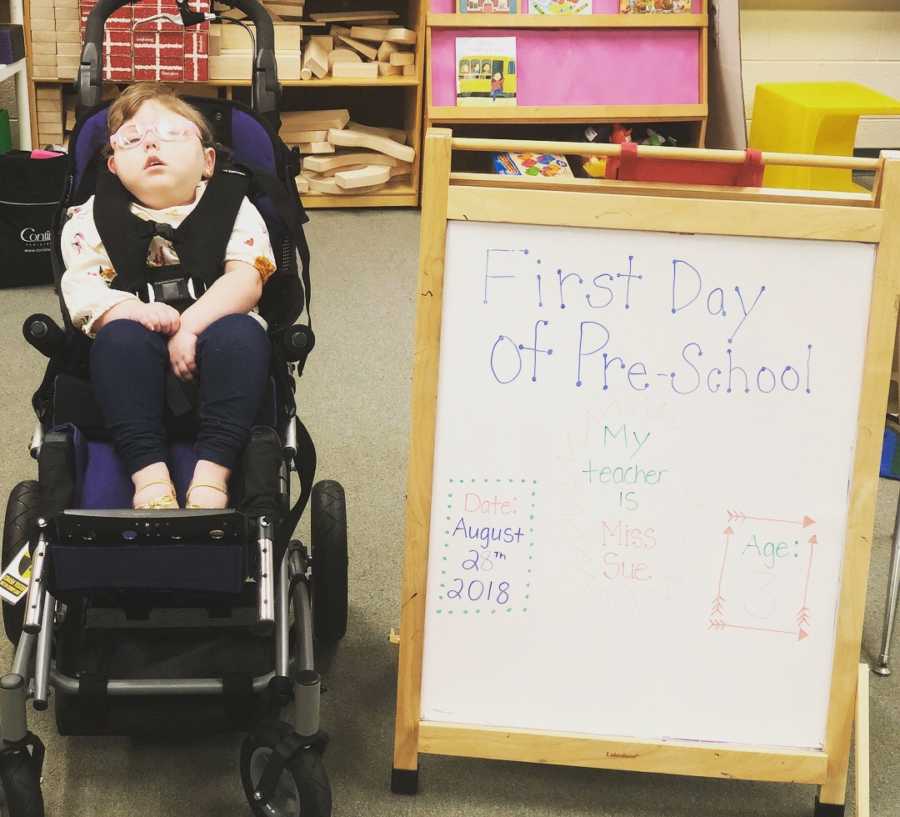 Little girl with bilateral open lip schizencephaly sits in wheelchair beside sign that says, "First Day of Pre-School"