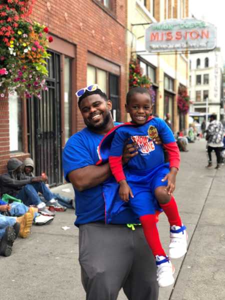 Father smiles as he holds up son dressed as super hero beside homeless people