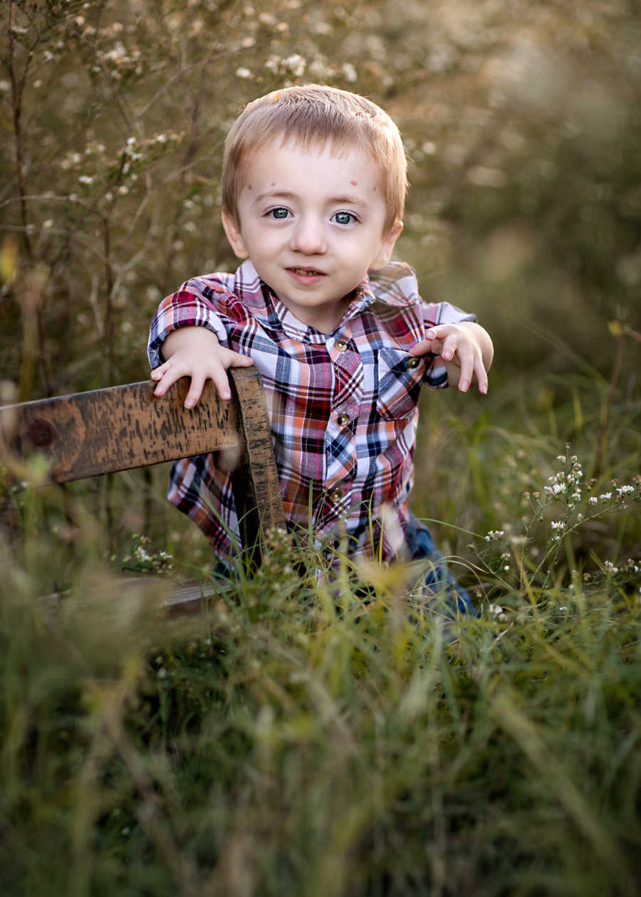Little boy with dwarfism stands in field holing onto small wooden chair
