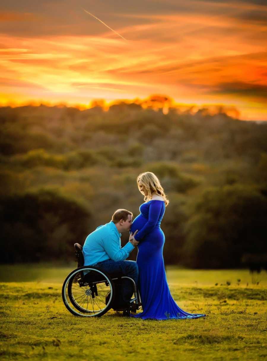 Pregnant woman stands while husband in wheelchair kisses her stomach in open field